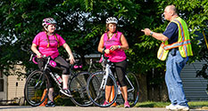Photo: 2 bicyclists in pink talk to guy in safety vest.